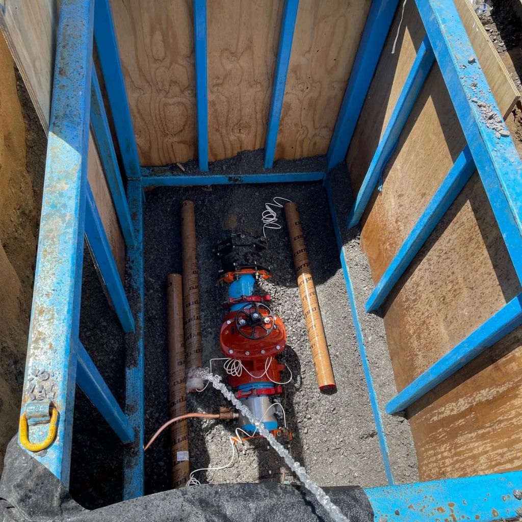 Vue en plongée d'une excavation de chantier sécurisée par un système d'étançonnement bleu conçu sur mesure. À l'intérieur, on observe un équipement de pompage orange et des tuyaux, indiquant probablement des travaux de maintenance ou d'installation souterrains.
