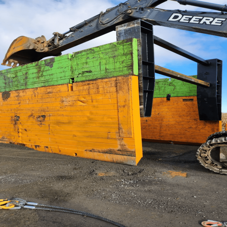 Une excavatrice Deere tient dans son godet un grand panneau de soutènement en bois composé de planches vertes et oranges, probablement pour une installation ou un retrait sur un chantier de construction. À l'avant-plan, on aperçoit une élingue en câble d'acier sur le sol. Un opérateur en gilet réfléchissant est visible en arrière-plan, assurant la sécurité du processus.