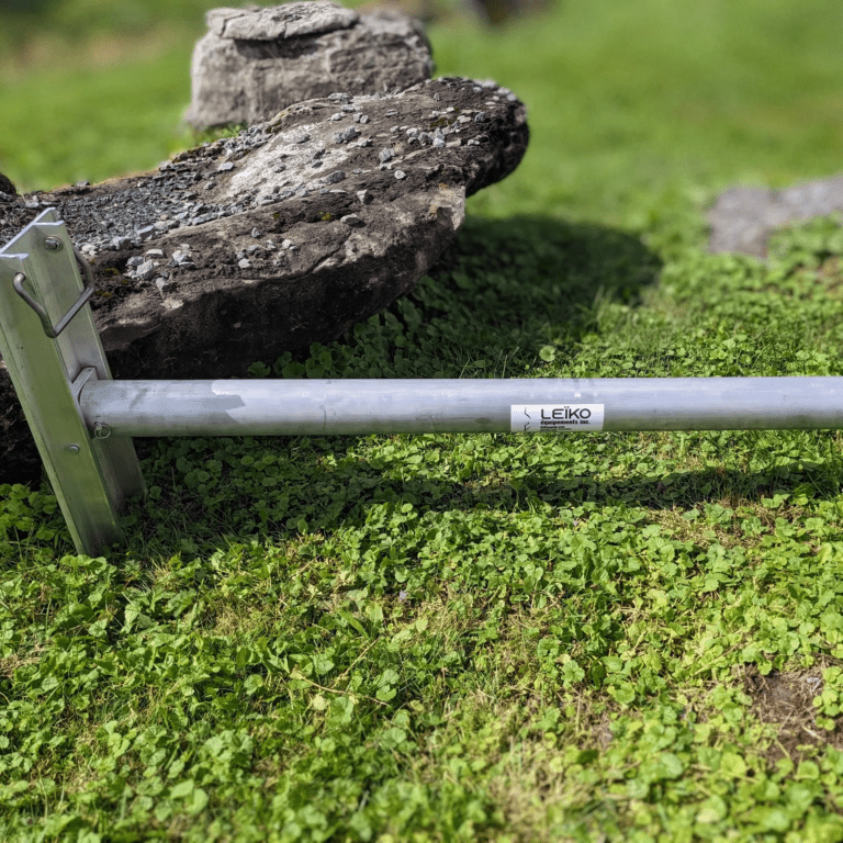 Bar de levage métallique de marque Leïko posé dans l'herbe verte, avec une partie attachée à une structure métallique en coin. À côté, une grosse pierre avec des lichens indique un environnement extérieur naturel.