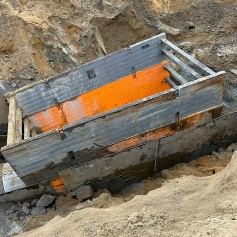 Caisson de blindage de tranchée déployé dans une excavation, avec des panneaux gris métalliques extérieurs et des renforts internes orange. La tranchée est entourée de terre érodée et de roches, indiquant un site de construction actif.