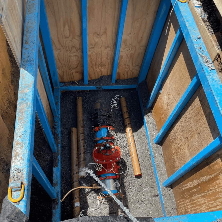 Vue en plongée d'une excavation de chantier sécurisée par un système d'étançonnement bleu conçu sur mesure. À l'intérieur, on observe un équipement de pompage orange et des tuyaux, indiquant probablement des travaux de maintenance ou d'installation souterrains.