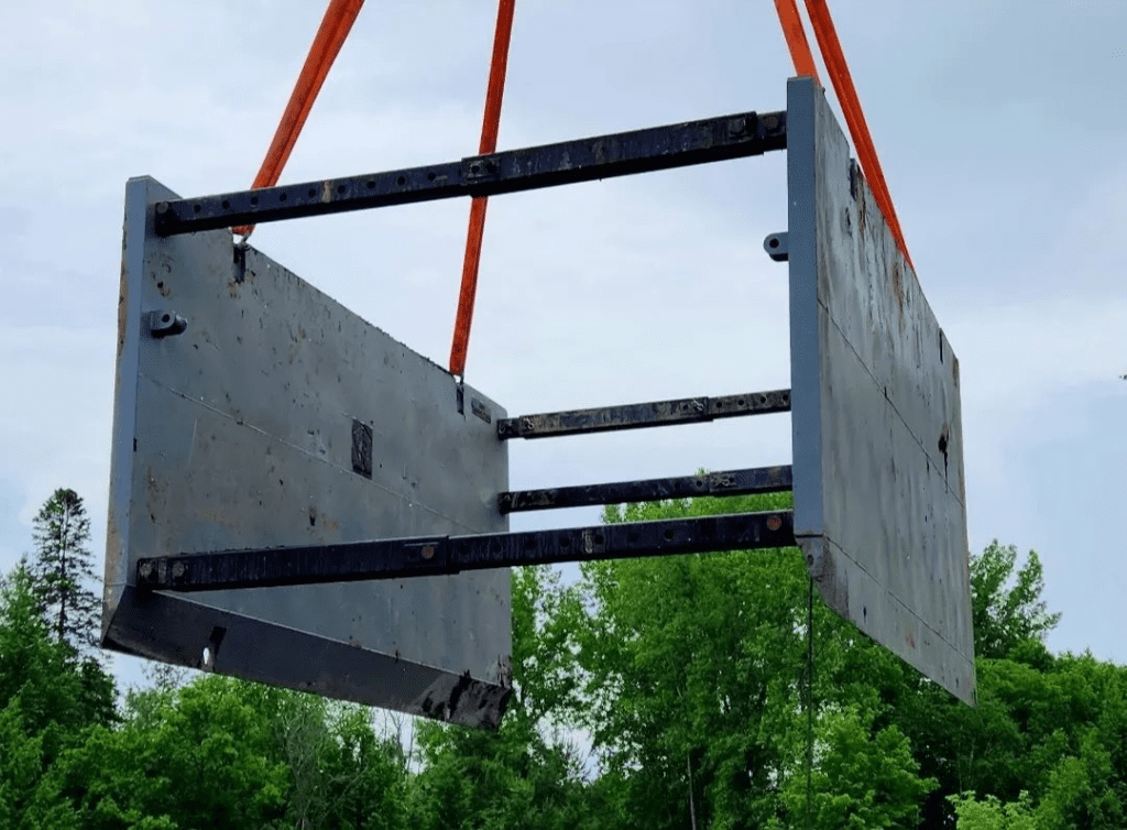 Un treillis métallique robuste de couleur grise, suspendu par des sangles de levage orange, destiné à la manutention de matériaux lourds sur un chantier de construction ou dans un environnement industriel.