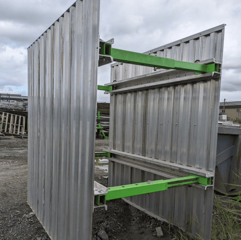 Porte métallique industrielle en position ouverte avec des barres de renforcement peintes en vert, installée sur un terrain de chantier avec un sol boueux, servant de barrière ou d'accès sécurisé à une zone de travail.