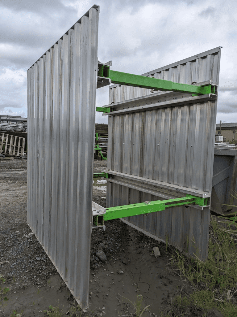 Porte métallique industrielle en position ouverte avec des barres de renforcement peintes en vert, installée sur un terrain de chantier avec un sol boueux, servant de barrière ou d'accès sécurisé à une zone de travail.