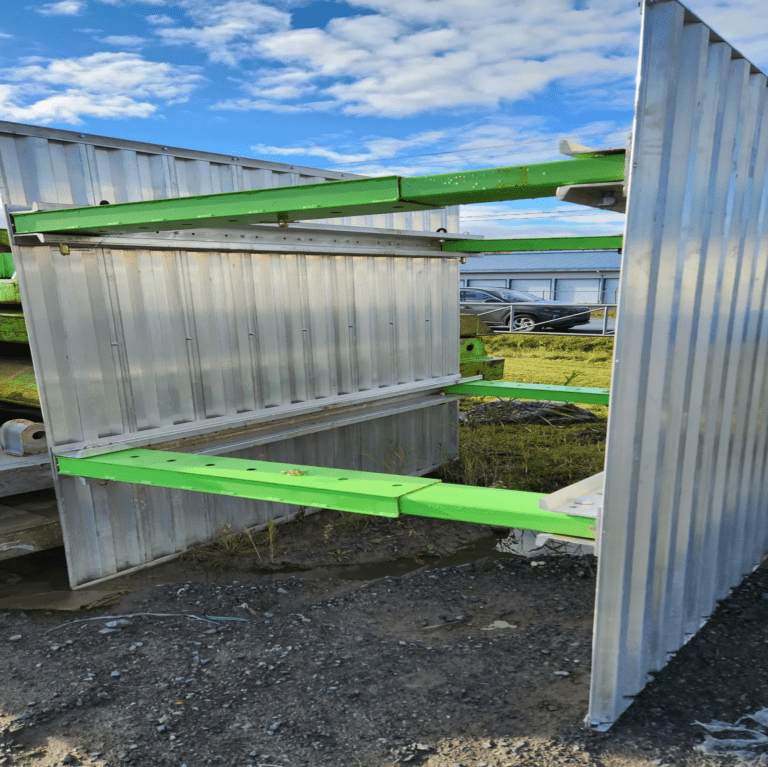 Vue latérale d'une porte de sécurité métallique semi-ouverte avec des renforts horizontaux verts, située sur un site de construction, montrant l'interaction entre la porte et les structures de soutènement environnantes.