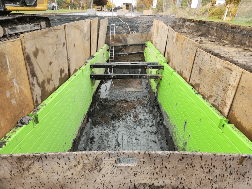 Installation de soutènement de tranchée en cours sur un chantier. Des panneaux de soutènement modulaires verts vifs sont en place dans une excavation profonde. Le chantier est situé près d'un plan d'eau, avec des sacs de sable formant une barrière en arrière-plan et du matériel de construction à proximité.