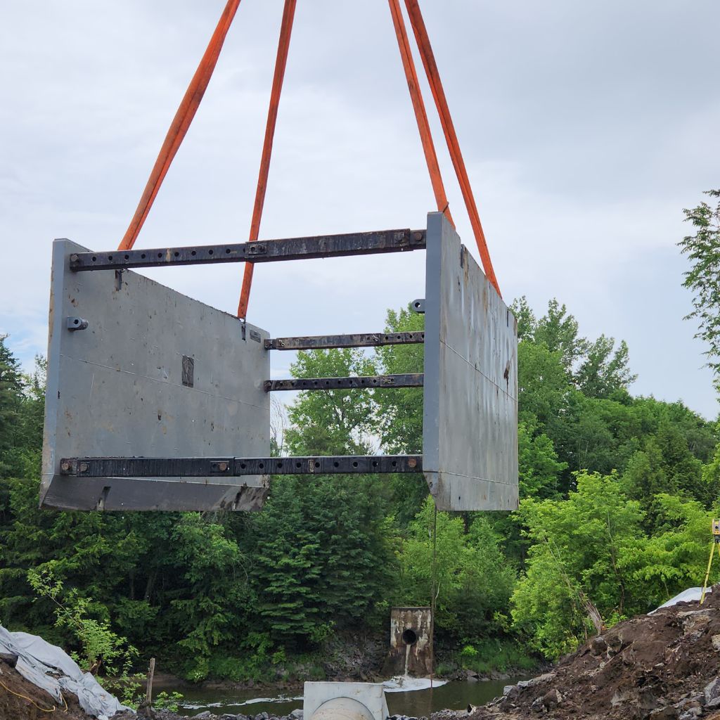 Un treillis métallique robuste de couleur grise, suspendu par des sangles de levage orange, destiné à la manutention de matériaux lourds sur un chantier de construction ou dans un environnement industriel.